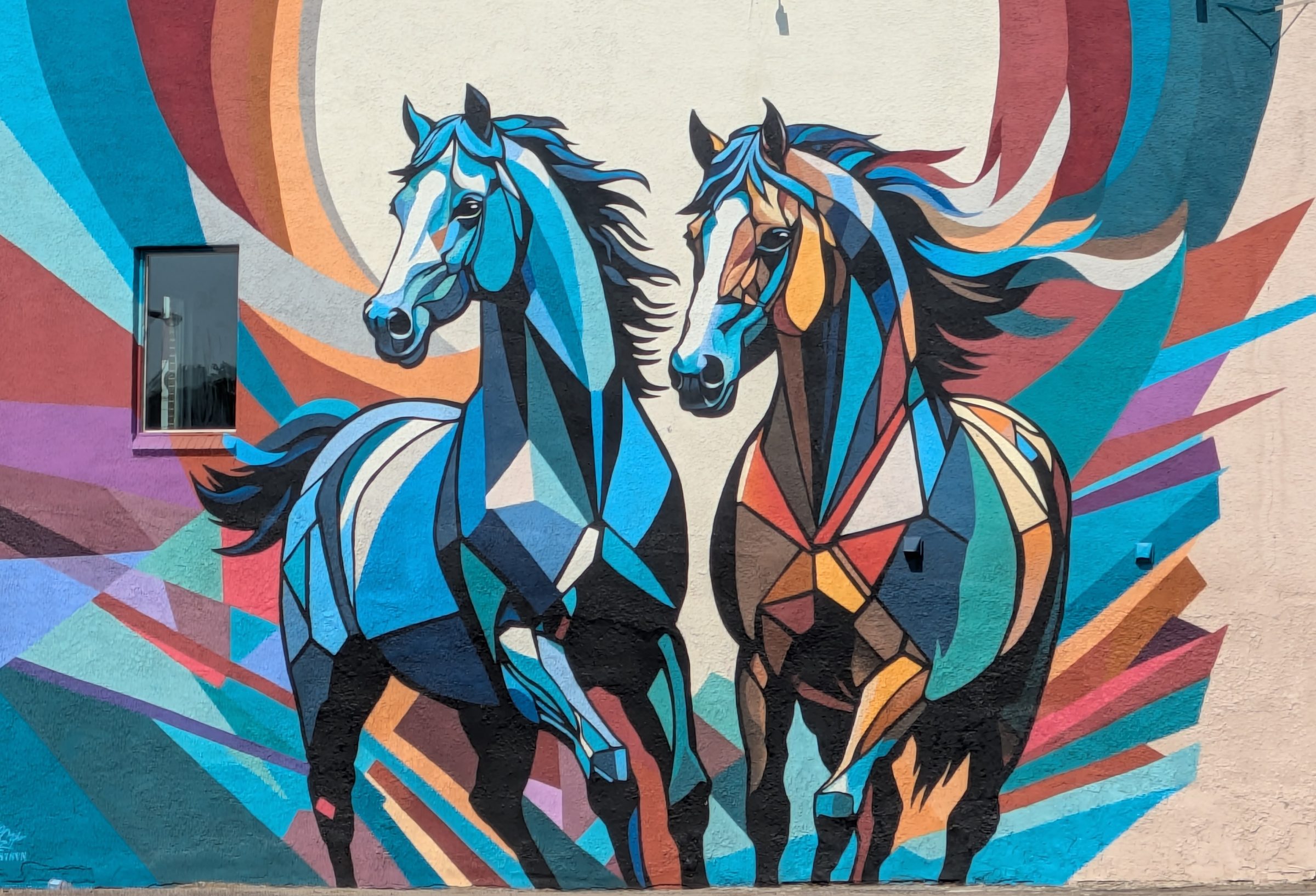 A geometric mural of two horses in blue, teal, rust, and orange, painted on an exterior wall.